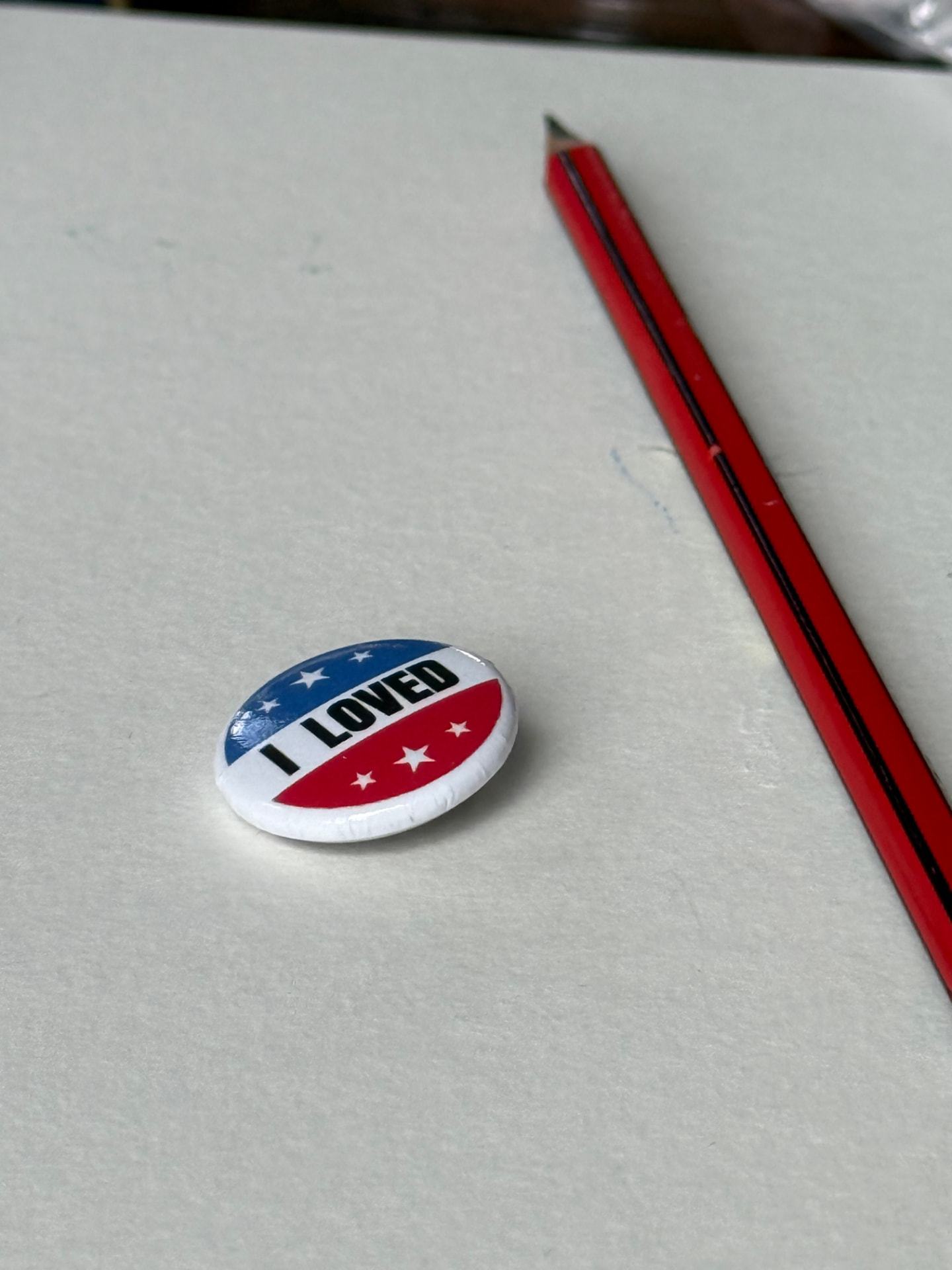 The round red, white and blue I LOVED button pin lying on white paper next to a red pencil, showing it is about one inch across